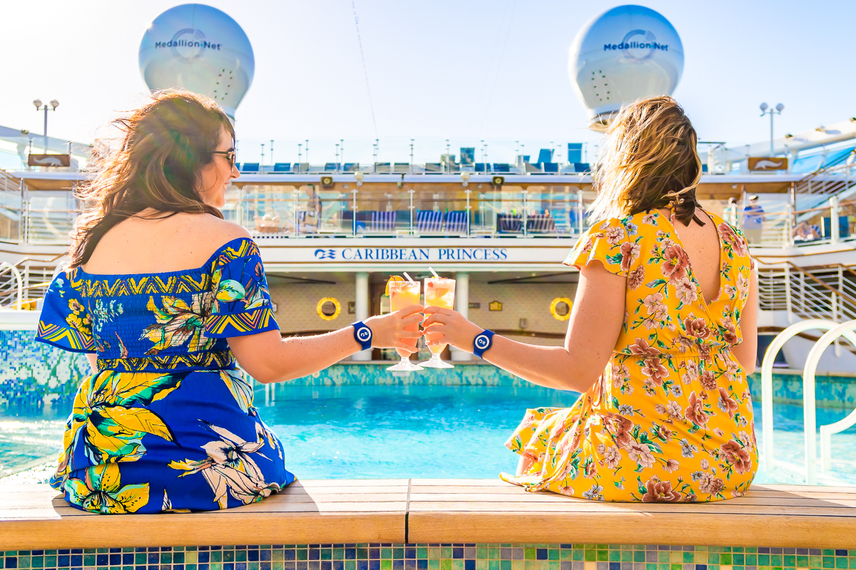 Two women cheers drinks sitting next to the pool on a cruise ship wearing floral dresses.