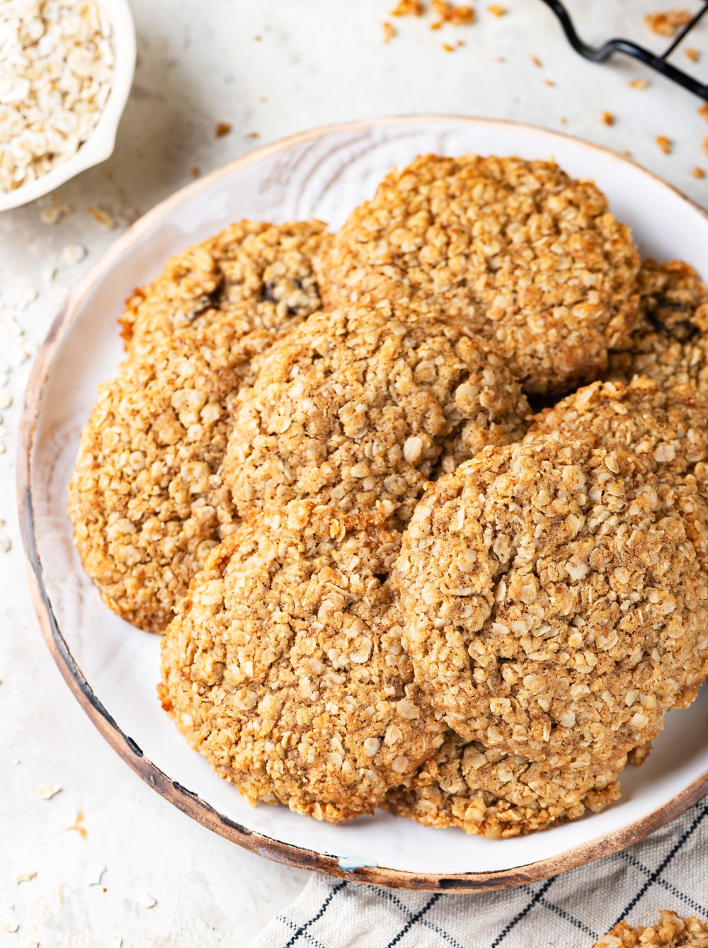 A white plate filled with eight vegan oatmeal cookies overlapping each other. The plate is on a white counter.