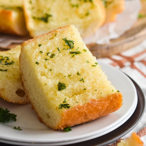 garlic bread slice on a white plate with fresh parsley on top and more garlic bread in the background