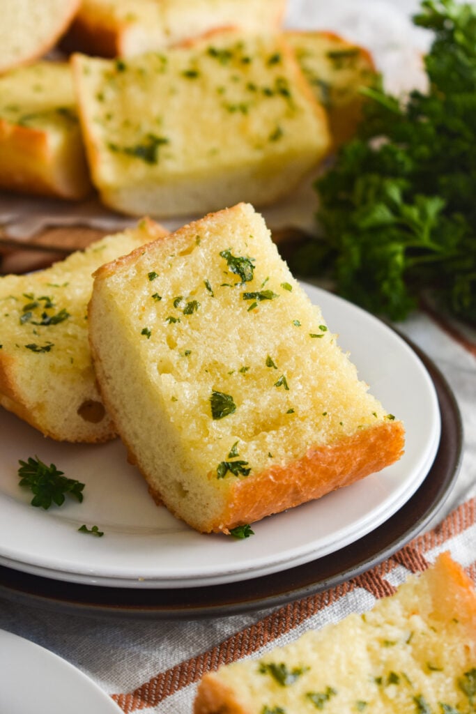 Slice of garlic bread on a white plate with more garlic bread and parsley in the background