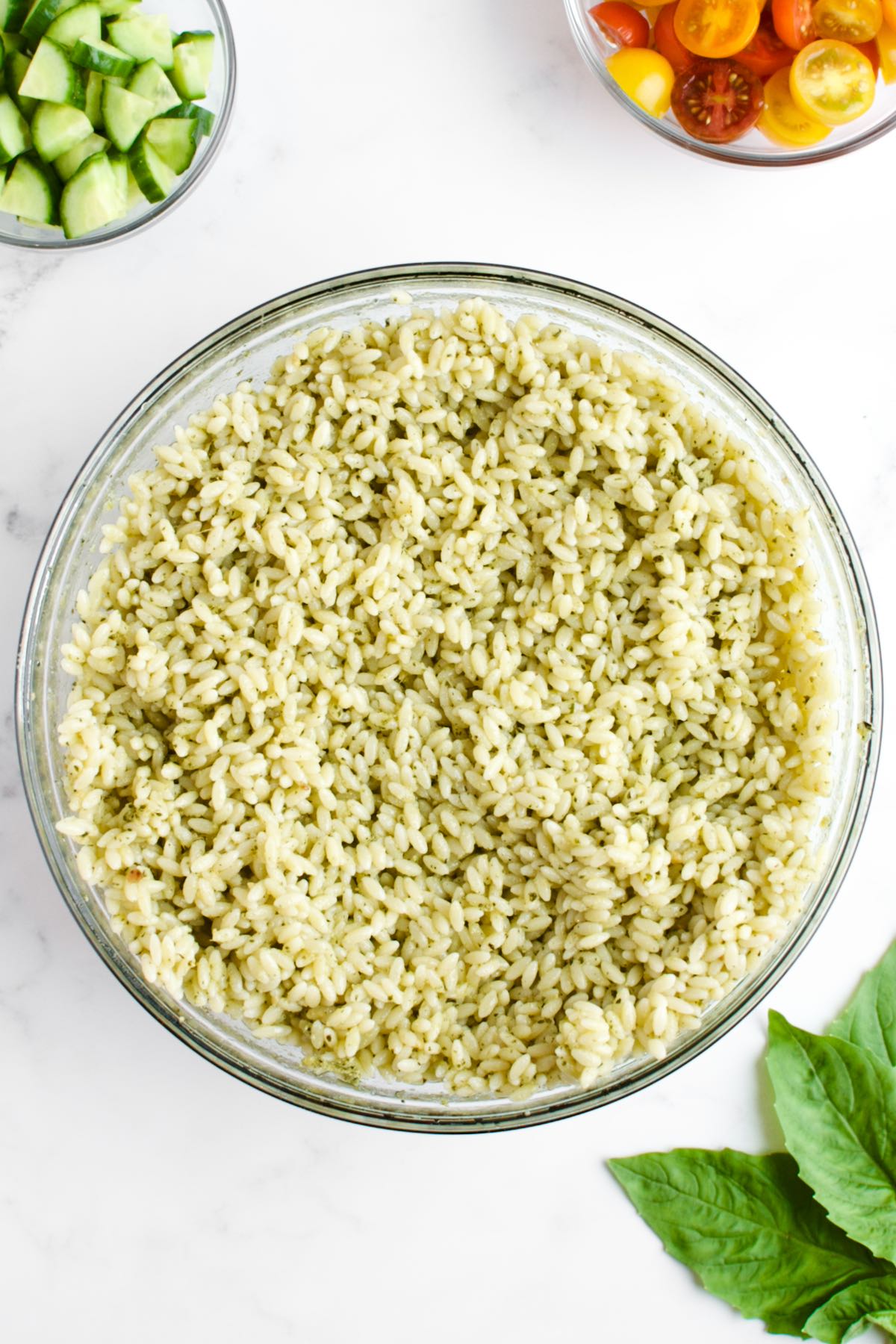 A glass bowl with cooked orzo with pesto dressing on a marble background.