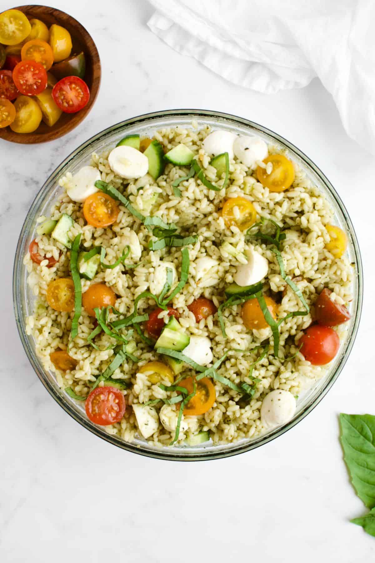 Orzo pesto pasta salad in a glass bowl on a marble background. Leaves of basil and a dish of cherry tomatoes are to the side.
