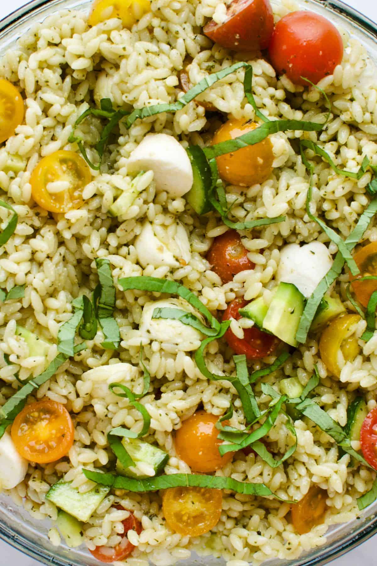 A close up view of orzo pesto pasta salad, with cherry tomatoes, cucumbers and basil.