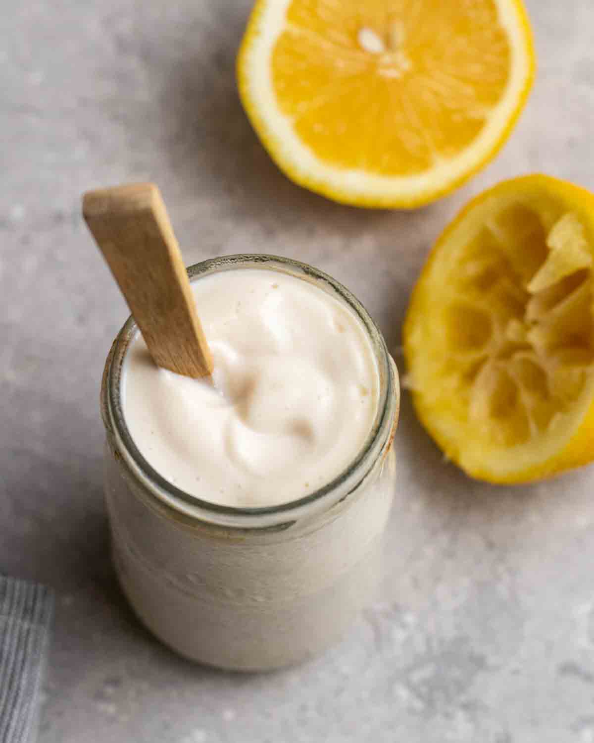 A jar of vegan sour cream with a wooden spoon beside with 2 lemon halves beside.