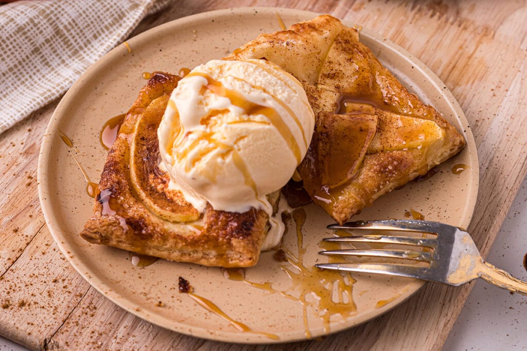 Round plate with a fork and puff pastry apple tart. A scoop of ice cream and caramel are on top of the tart.