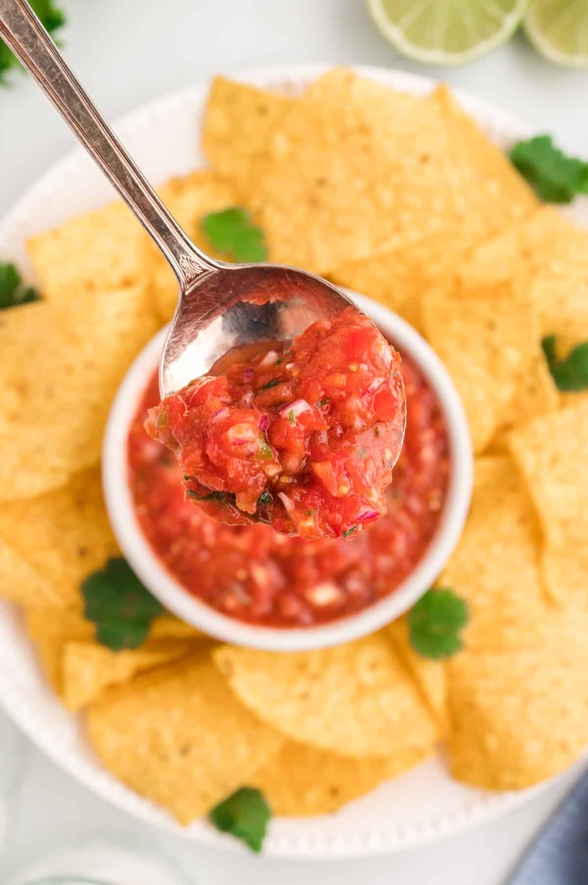 A bowl of salsa with a spoon scooping salsa out. A platter of tortilla chips is in the background.