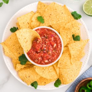A bowl of salsa on a white platter, with tortilla chips surrounding it. One chip is in the salsa with a scoop of salsa on it.