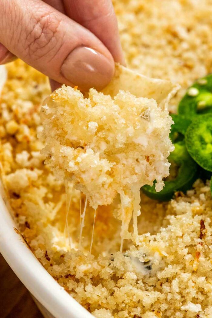 Close up side view of a hand digging a curved chip into jalapeno popper dip, lifting up a bite, with stringy cheese dripping back into the dish.