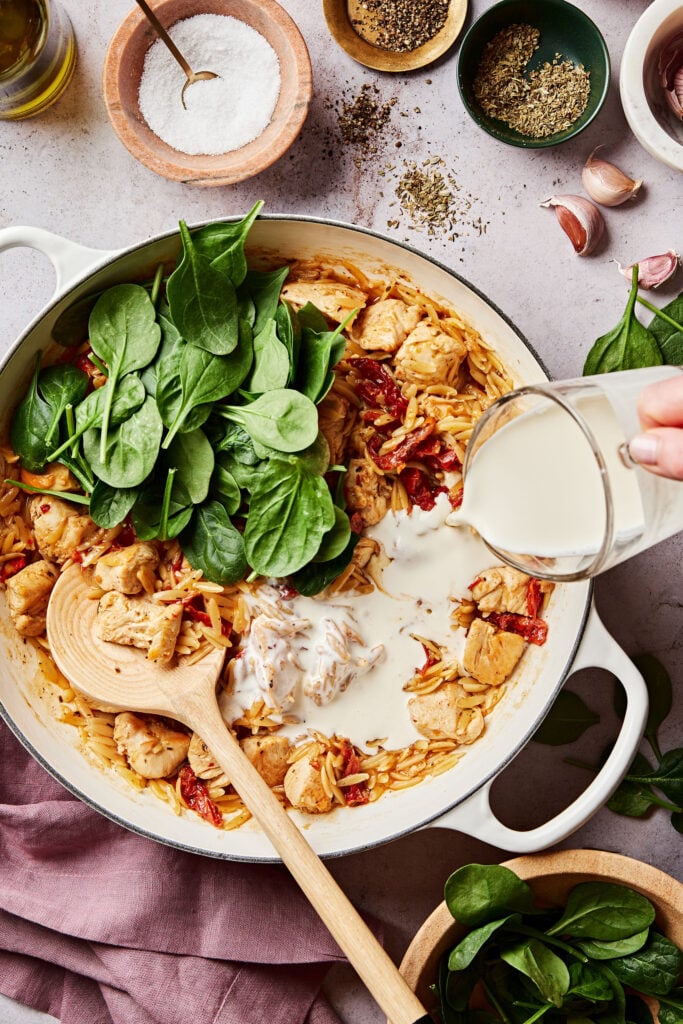 A pan of the one pot marry me chicken orzo, with the fresh spinach and non-dairy cream actively being poured into it 