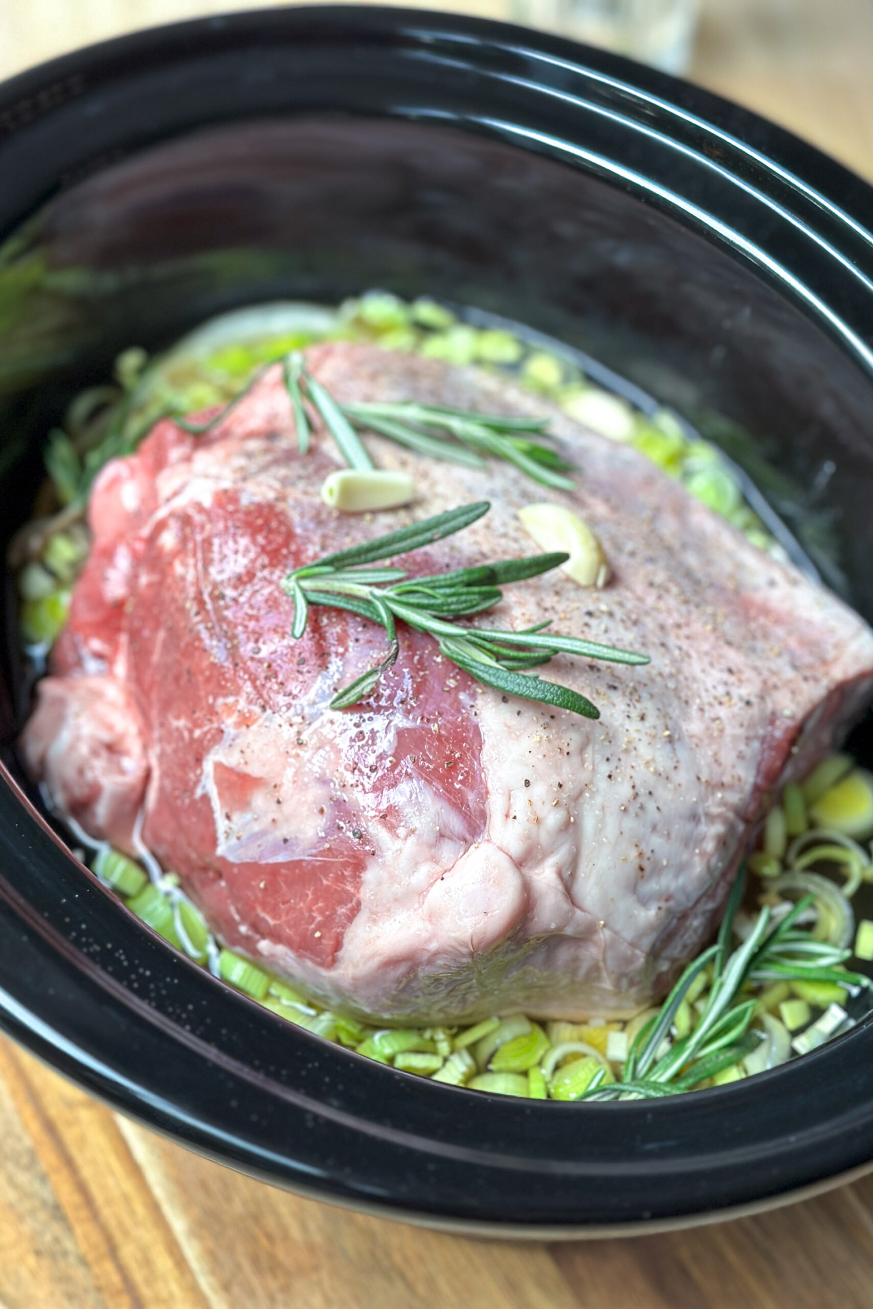 Side view of lamb roast with fresh rosemary in crockpot insert.
