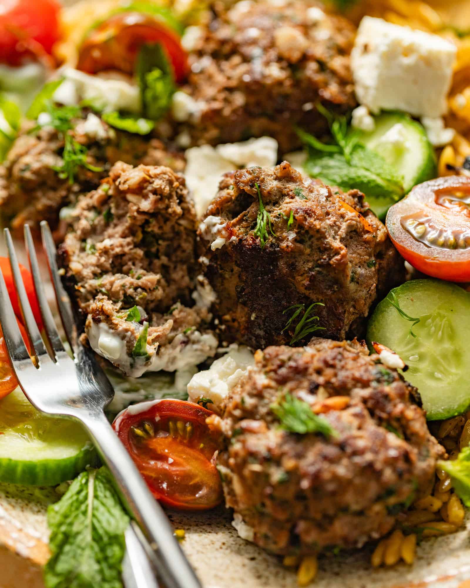 up close photo of greek meatballs in a bowl with a fork and blocks of feta cheese and tomatoes.