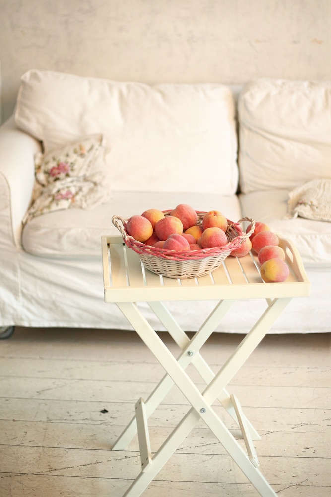 A neutral white couch with a chabby-chic style throw pillow behind a simple off white tray table holding a white backset with two handles and a dark pink rim holding peaches.
