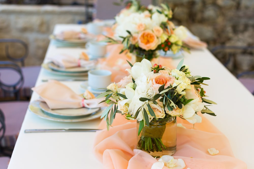A white linen tablecloth covers a table with light blue dishes and peach colored napkins. There are simple peach, white, and green bouquets in glass vases.