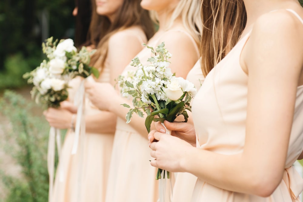 Side view of bridesmaids in peach fuzz colored strappy gowns holding simple white flowers tied with a white bow.