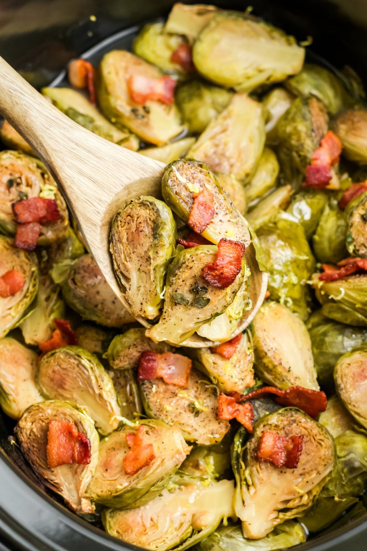 A wooden spoon lifting some cooked Brussels sprouts up from the slow cooker.