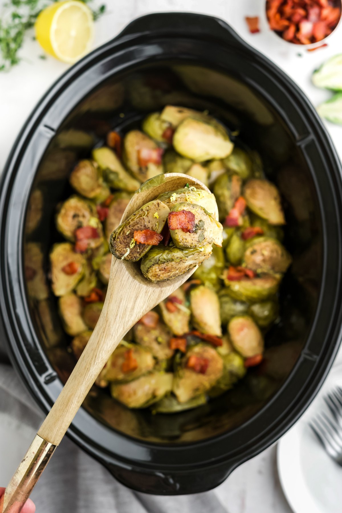 A wooden spoon holding up a serving of Brussels sprouts with bacon over the crockpot.