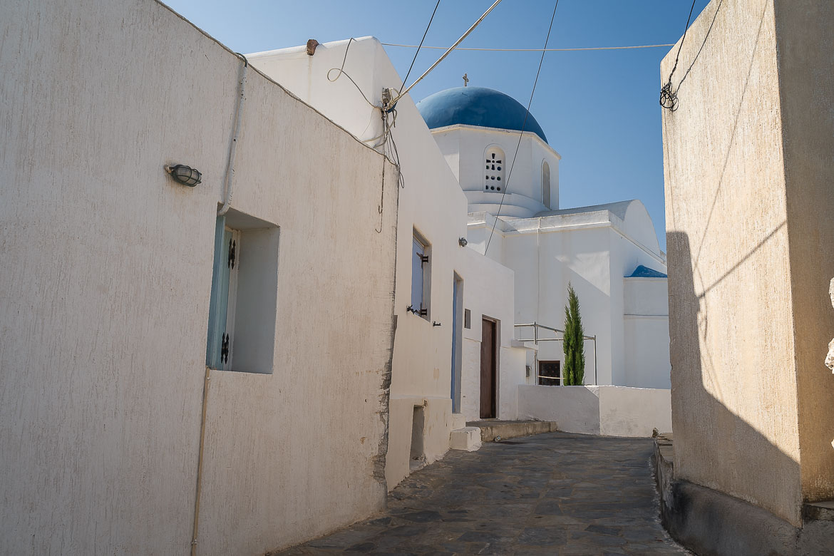 A narrow paved street with whitewashed buildings and a blue domed church in Panagia Village.