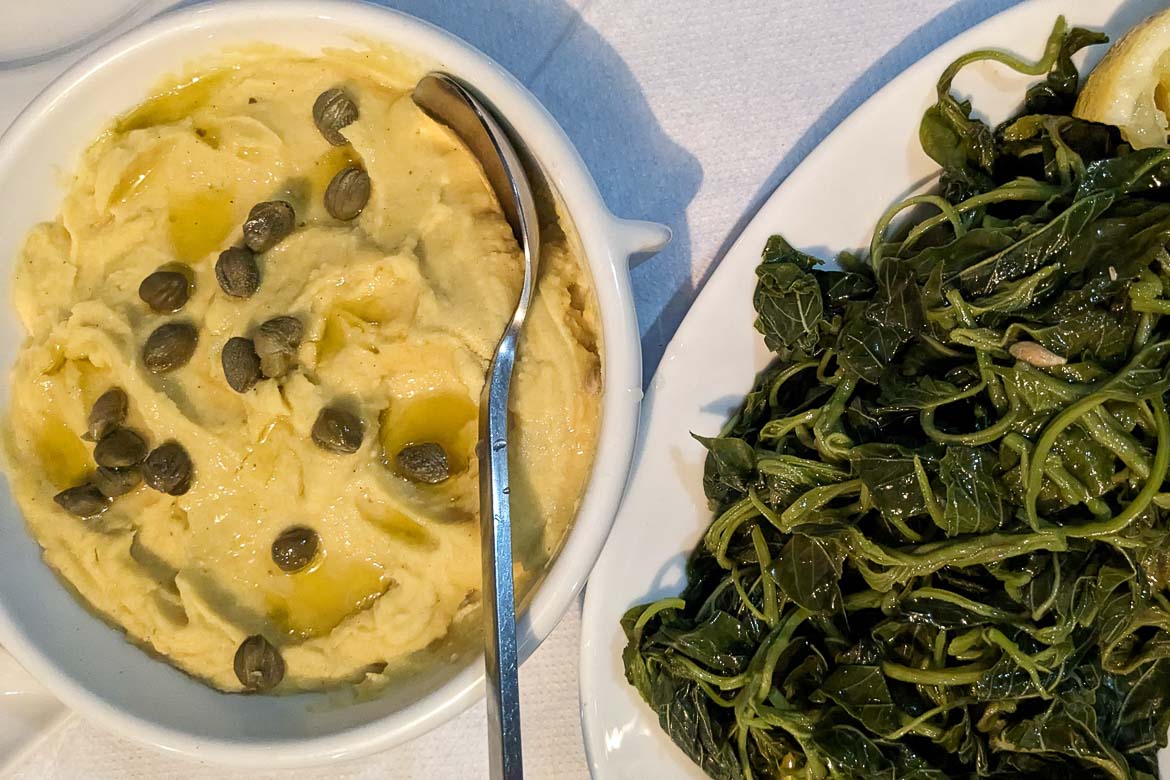 A fava puree dish with capers and olive oil on top. Next to it, there is a dish of boiled greens with lemon.