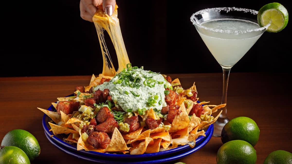 A hand pulling a stretch of melted cheese from a loaded plate of nachos with a margarita on the side, showcasing Mexican foods that aren't from Mexico.