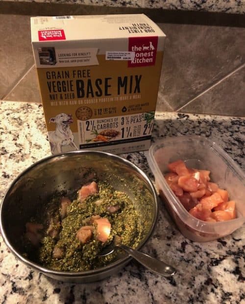 A package of THK's base mix is sitting on a granite kitchen counter behind a stainless steel dog bowl filled with homemade dog food and an open plastic counter with raw chicken breast.