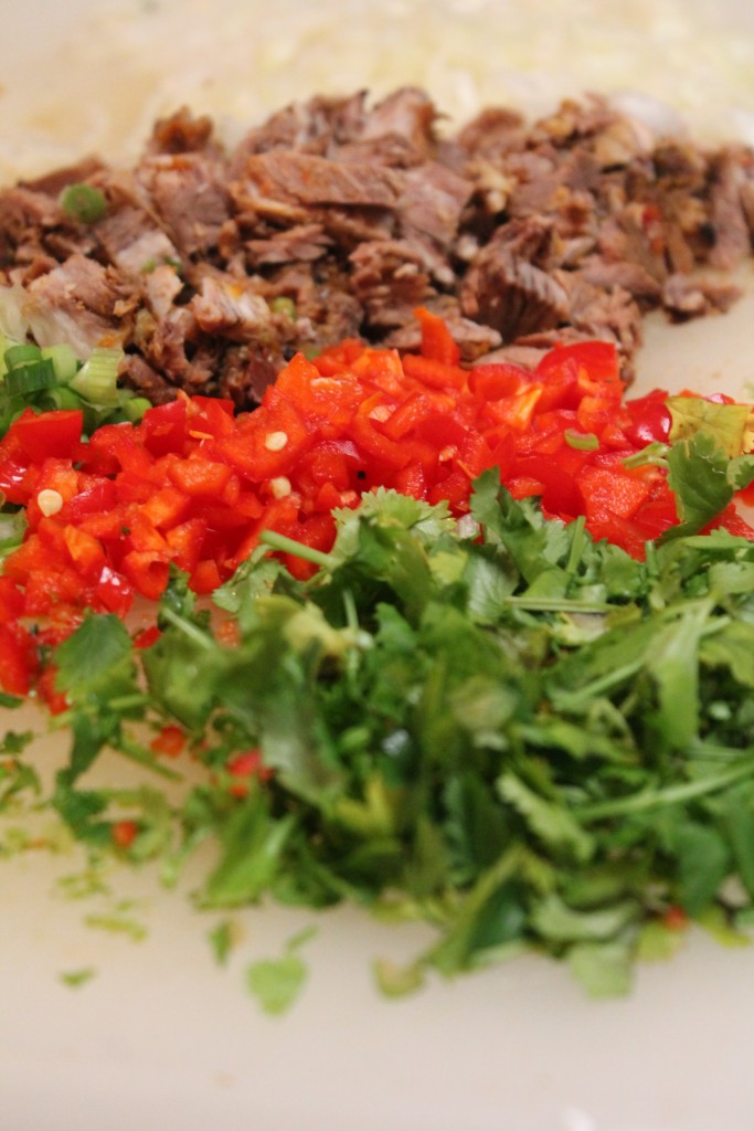 A closeup image of chopped brisket, red bell peppers, green onions, and fresh cilantro on a cutting board.