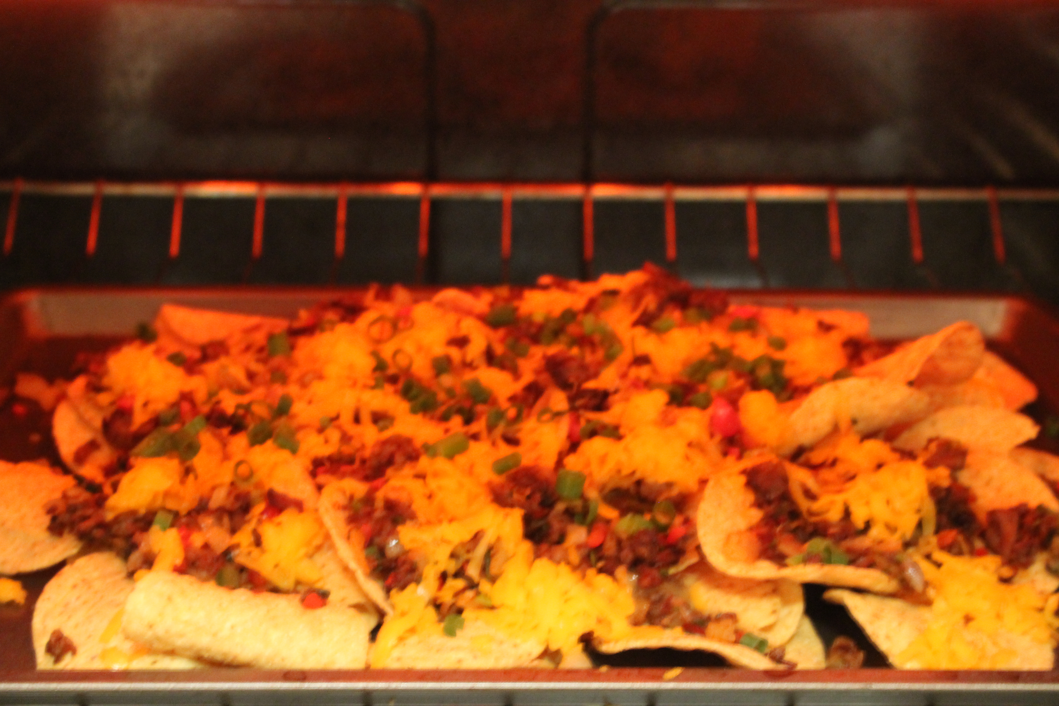 An image of a sheet pan of brisket nachos cooking in an oven until the cheese is melted and bubbling.