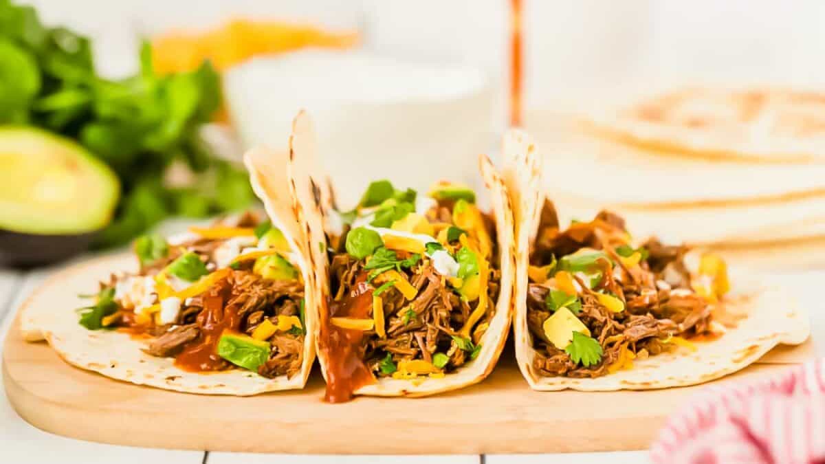 Three pulled beef tacos on a wooden cutting board.