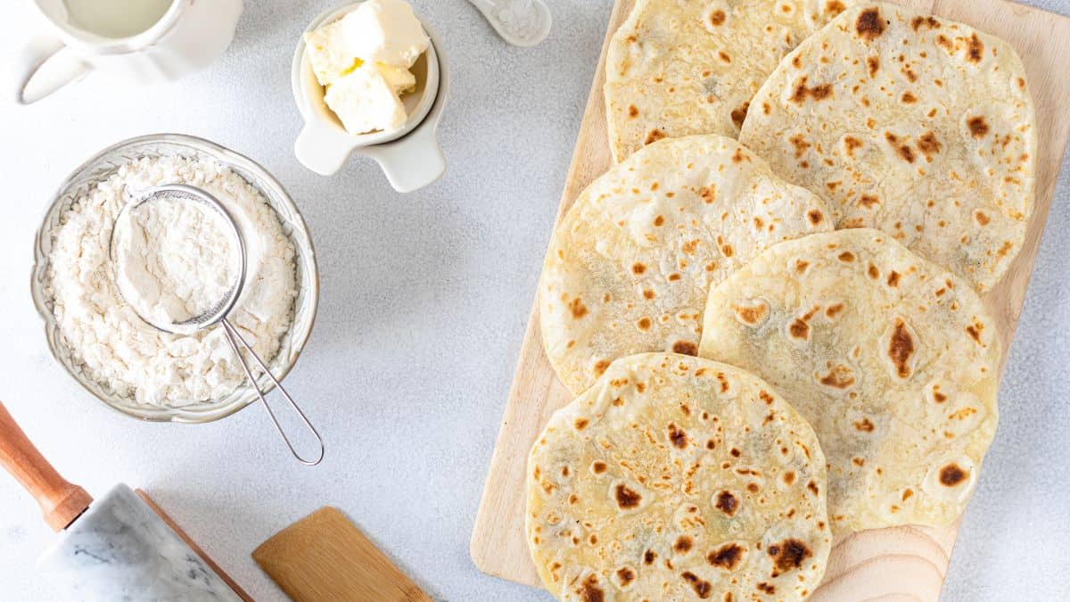 Fresh homemade flatbreads on a wooden board with ingredients and utensils around.