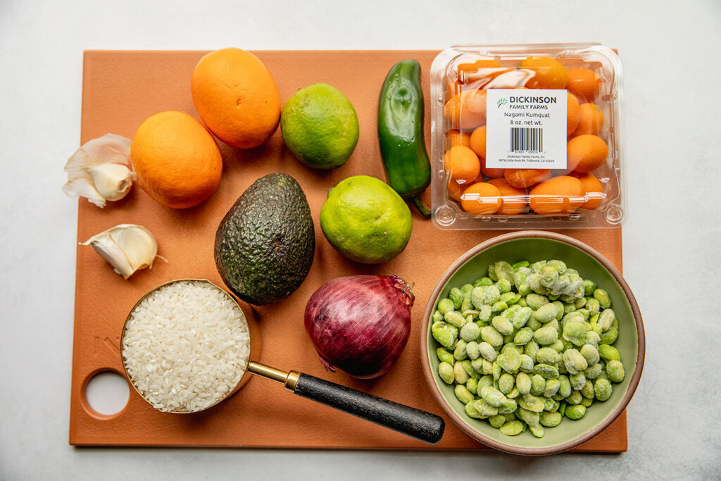 Cutting board topped with blood oranges, avocado, edamame, rice, red onion, lime, jalapeno, container of kumquats, and garlic.
