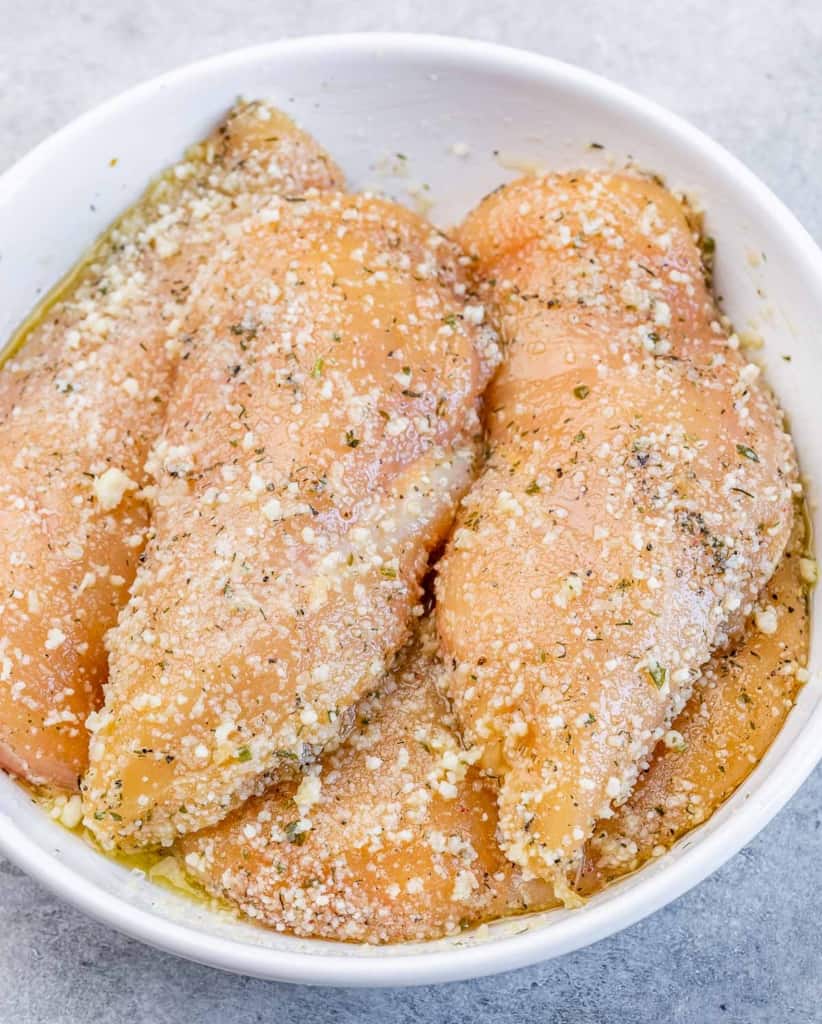 Raw chicken in a bowl covered with parmesan and seasonings.