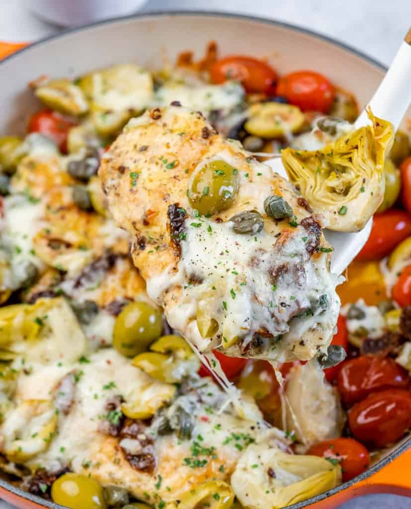 spatula grabbing a cooked chicken breast from skillet topped with melted cheese and olives with fresh tomatoes 