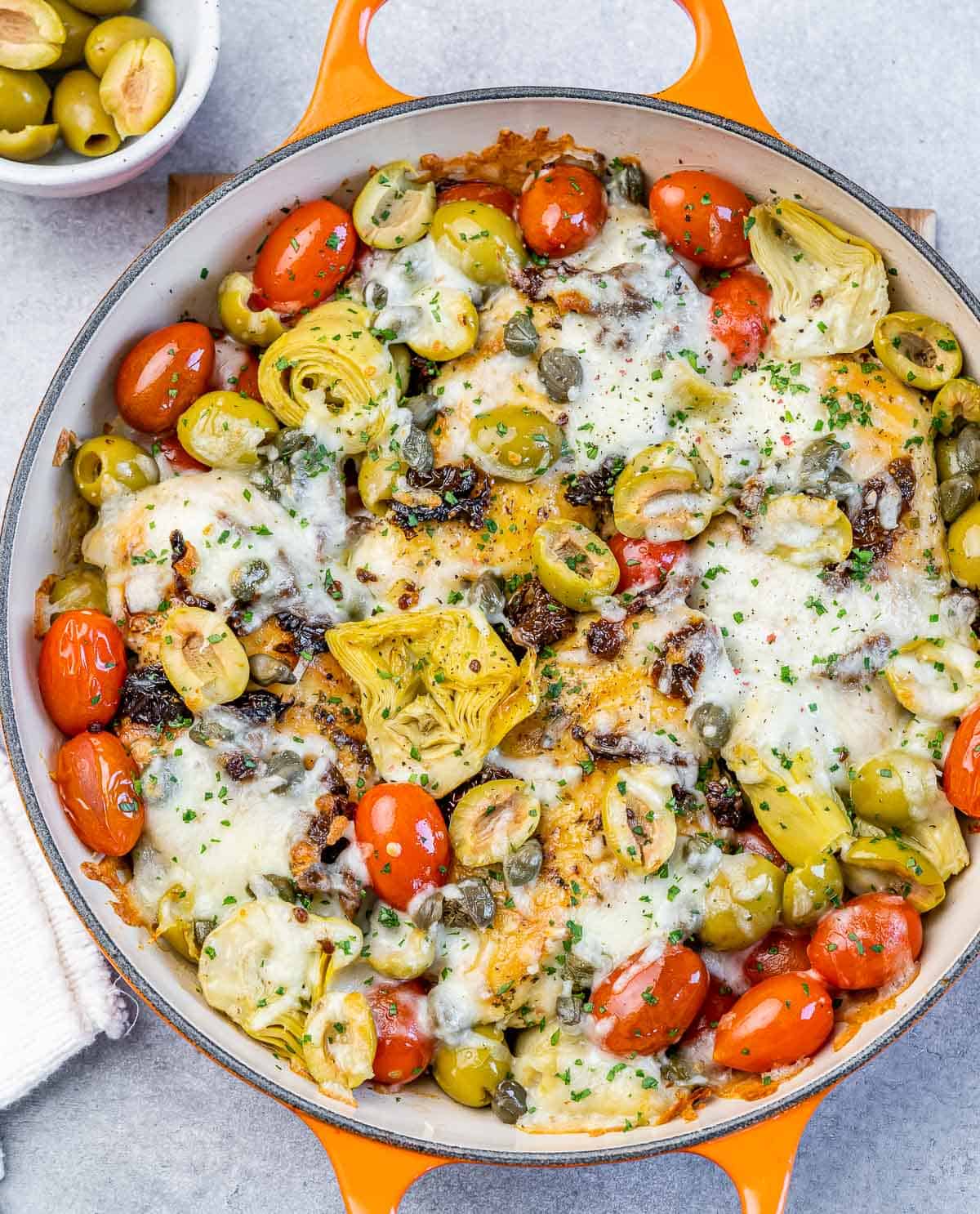 Baked Italian chicken in a skillet with tomatoes, olives, artichokes, capers and cheese.