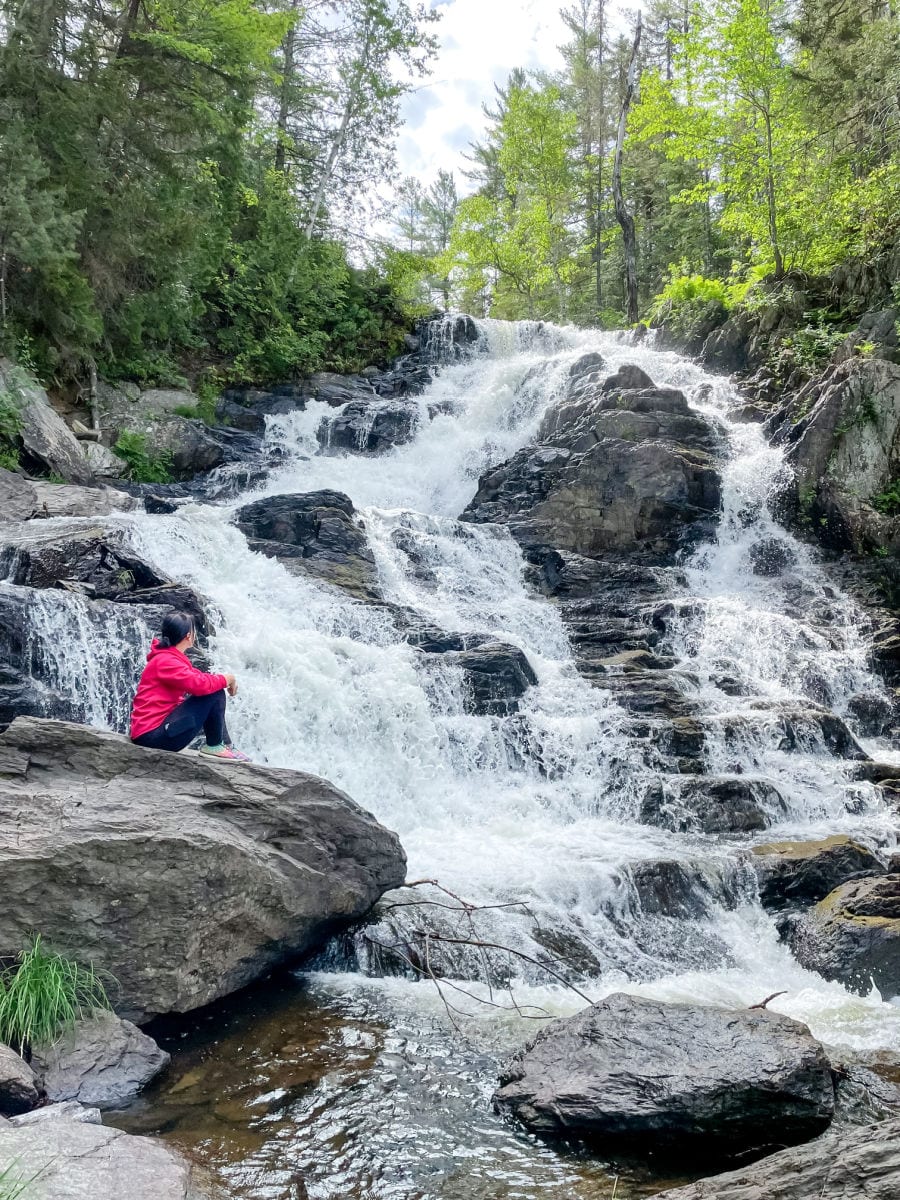woman in red jacket and black pants sitting on rocks looking at Shin Brook Falls