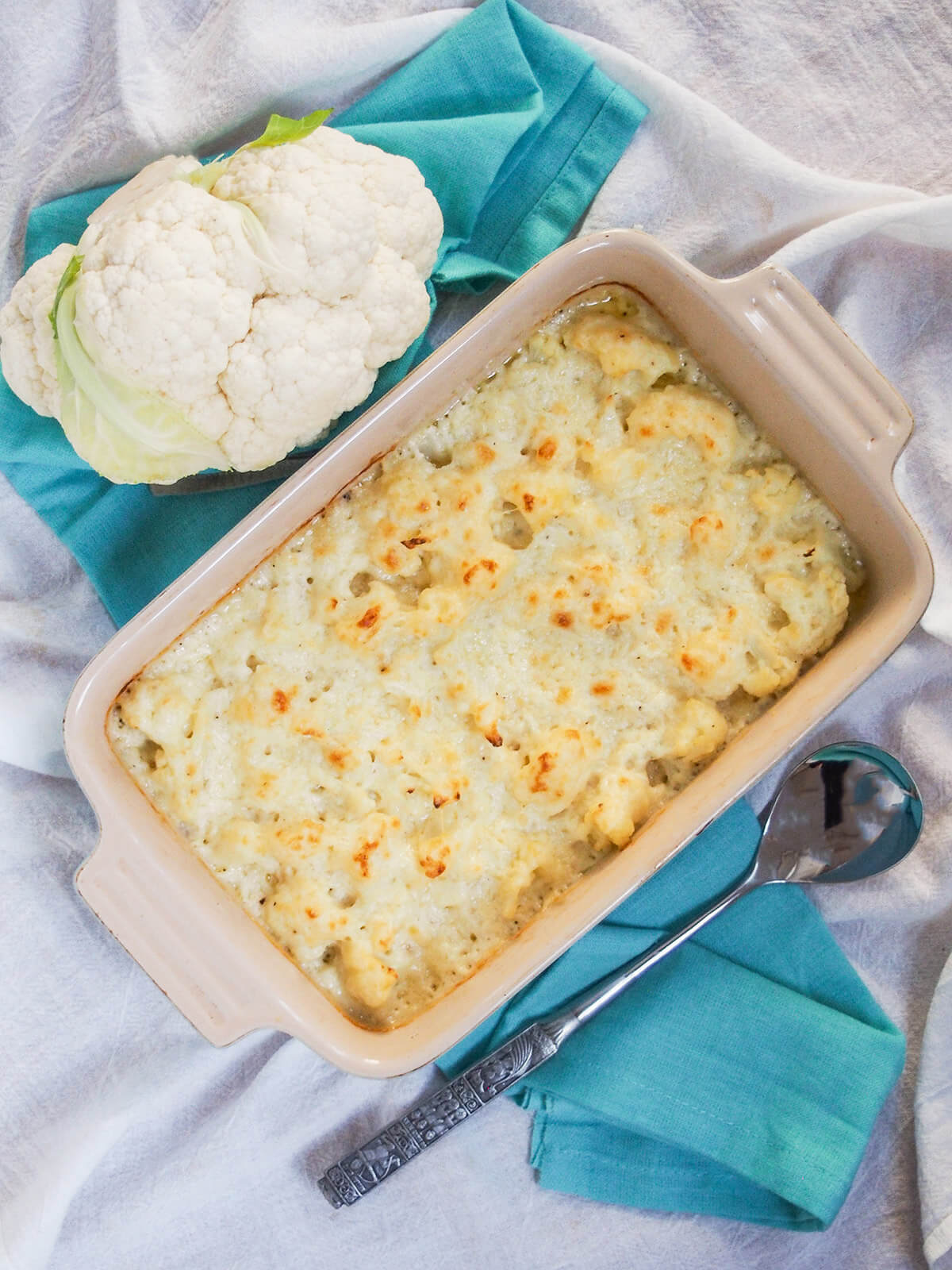 dish of cauliflower cheese with part caulifower to one side and serving spoon on other