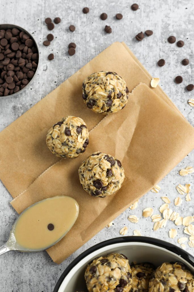 Tahini Chocolate Chip Bliss Balls on top of parchment paper with chocolate chips and oats scattered and spoon of tahini 