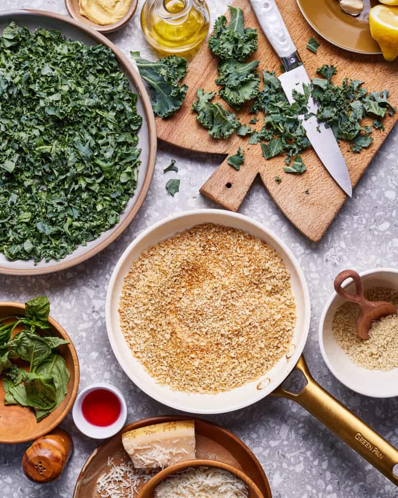 A bowl, filled with the parmesan cheese and breadcrumbs, sitting on a countertop next to a bowl of chopped kale 