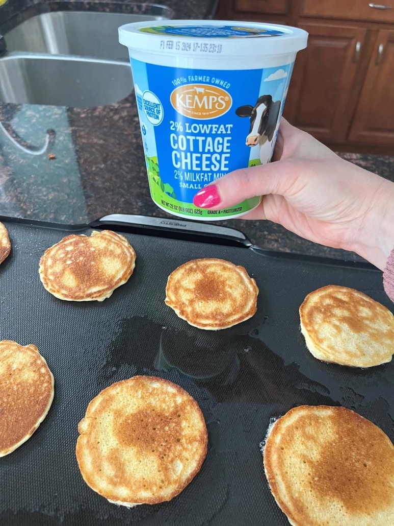hand holding a container of cottage cheese above a set of Cottage Cheese Pancakes on a griddle