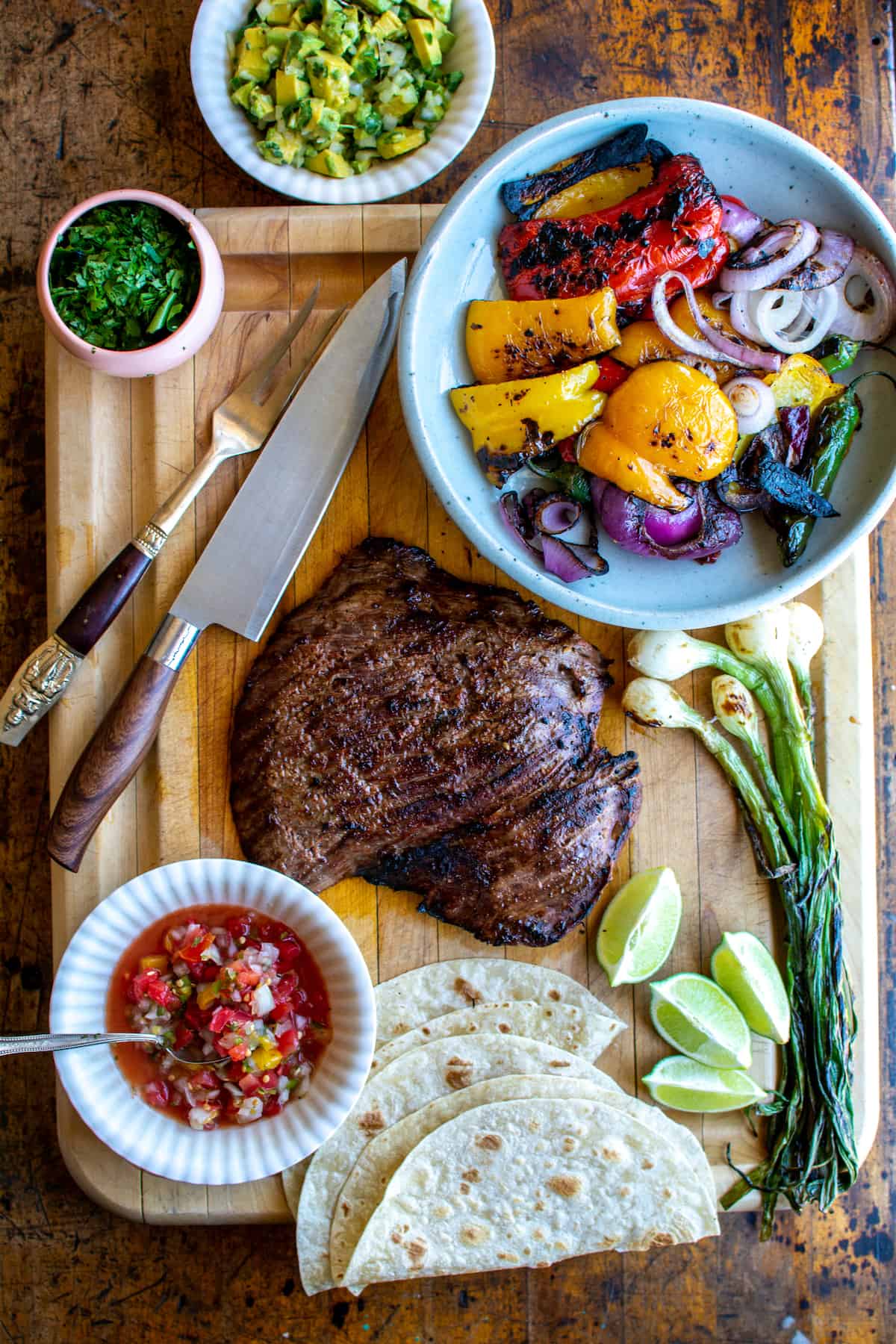 Steak fajitas on a wood cutting board surrounded by pico de Gallo, grilled peppers and onions, flour tortillas, and grilled onions