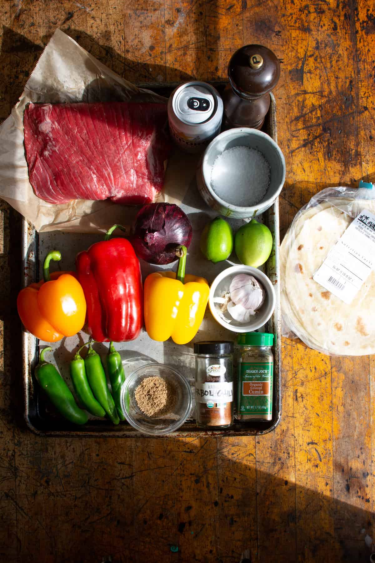 All the ingredients to make steak fajitas including flank steak, beer, pepper, salt, red onion, limes, bell peppers, and more. 