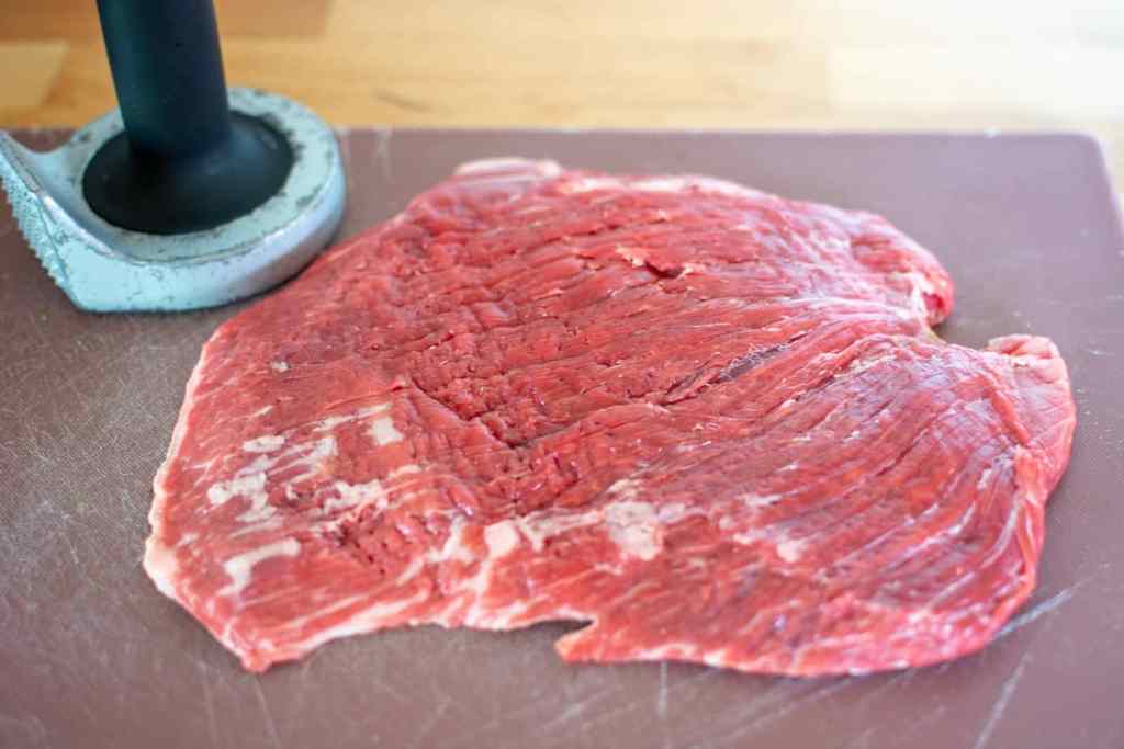 A piece of flank steak pounded thin on a red plastic cutting board with a meat mallet behind it. 