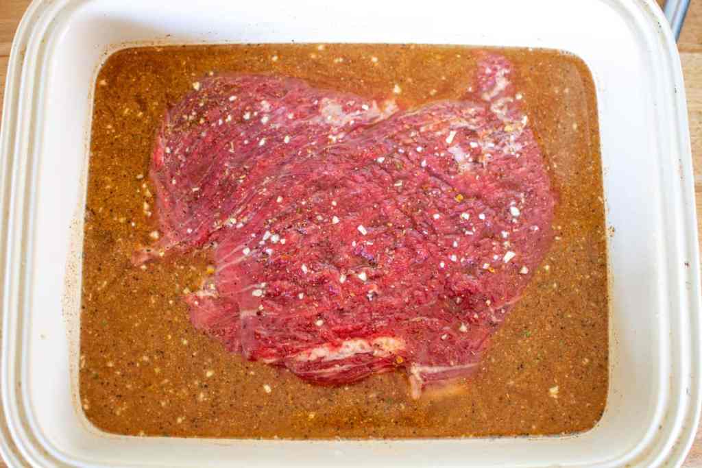 A piece of flank steak in fajita marinade in a ceramic baking dish on a wood counter. 