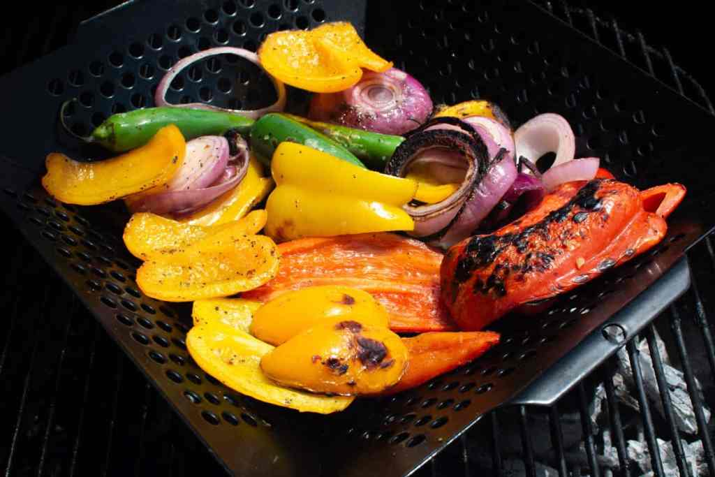 Grilled colorful bell peppers in a grill basket on the grill along with serranos, and red onion slices. 
