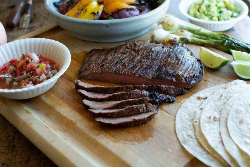 A flank steak sliced into strips on a wood cutting board next to some lime wedges, flour tortillas, grilled bell peppers, and more.