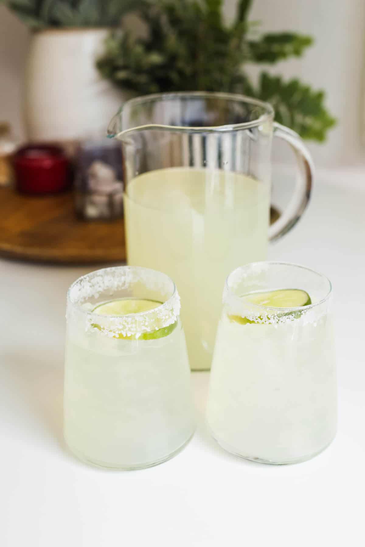 A pitcher of margaritas on a table with two salt rimmed glasses in front of it with margaritas.