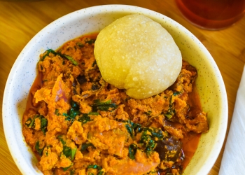 stone bowl of Nigerian efo elegusi (soup) served with a ball of eba swallow next to a cold glass of hibiscus zobo drink.