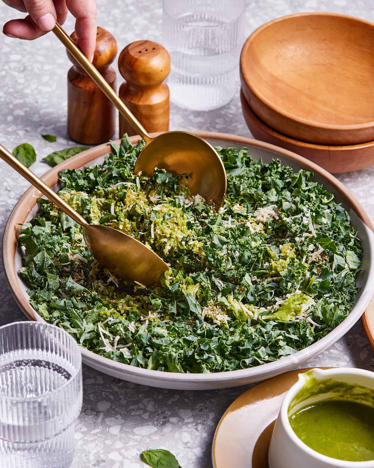 A bowl of the kale salad with lemon-basil dressing, sitting on a countertop with metal serving utensils resting in the bowl