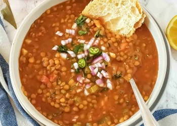 A soup bowl filled with homestyle lentil soup with vegetables, served with baguette, chopped onions, cilantro leaves and lemon wedges.