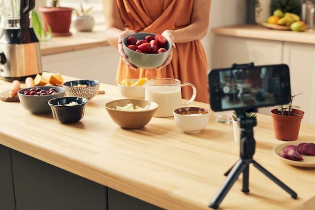 A woman cooking while recording a video with a phone on a tripod.