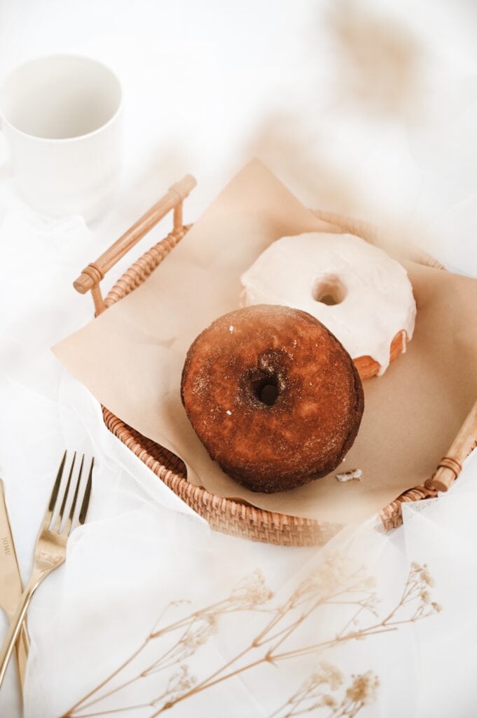two air fryer biscuit donuts served in a wooden basket, one topped with white chocolate glaze and the other is topped with cinnamon and powdered sugar