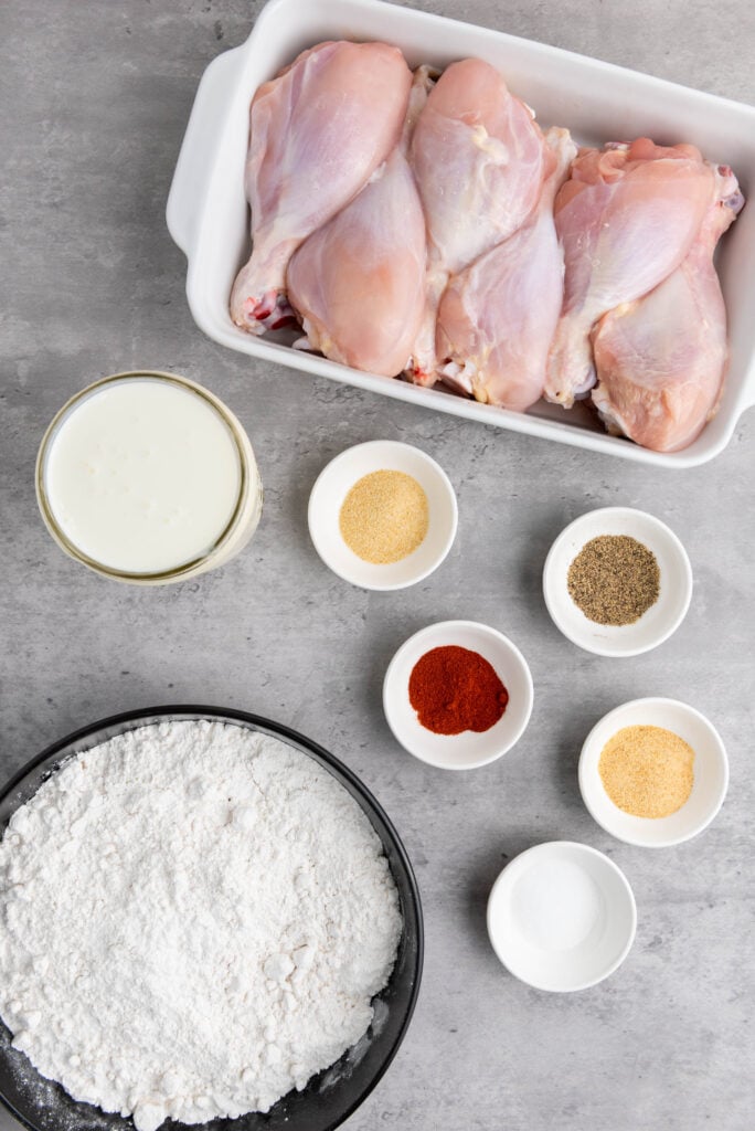 Ingredients for  preparation: raw chicken breasts, flour, spices, and a bowl of buttermilk batter on a kitchen surface.
